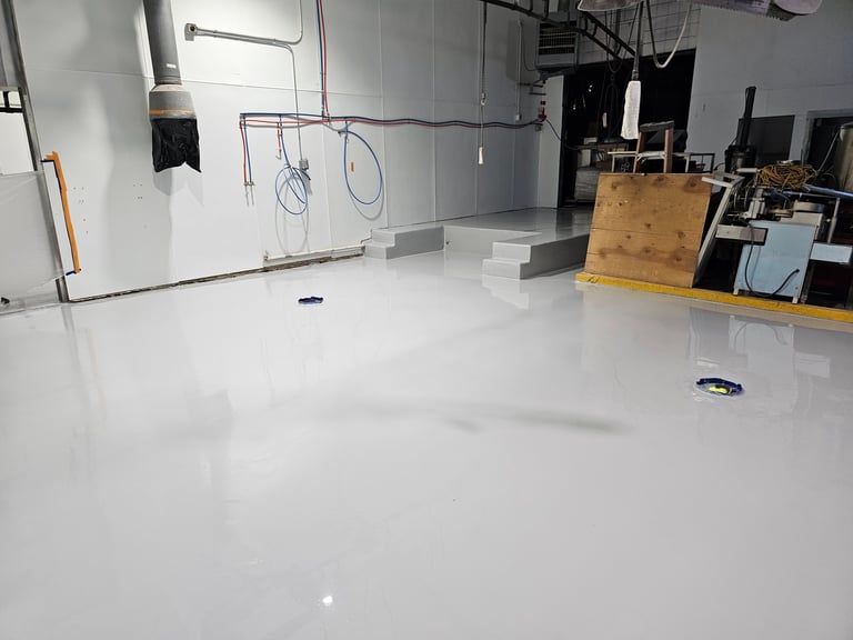 Empty industrial warehouse or studio space with polished white epoxy floor, overhead wiring, and scattered equipment and wooden crates