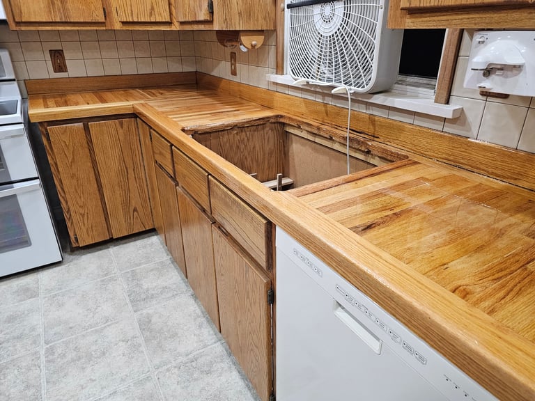 Kitchen under renovation with wooden cabinets and countertops, sink removed, appliances visible in background
