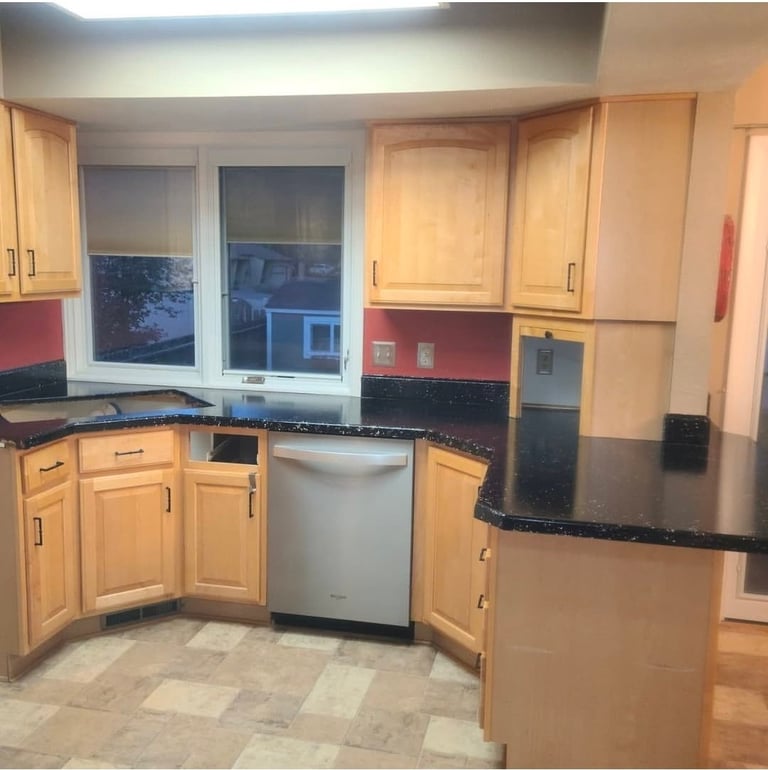 Modern kitchen with light wood cabinetry, black granite countertops, stainless steel dishwasher, and windows overlooking a residential neighborhood