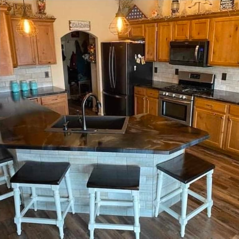 Warm-toned kitchen with wood cabinets, dark granite island with white base and two black stools, stainless steel appliances, subway tile backsplash, and hardwood flooring