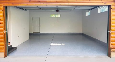 Empty garage interior with open double doors, gray concrete floor, white walls, and wooden log exterior siding visible at frame edges